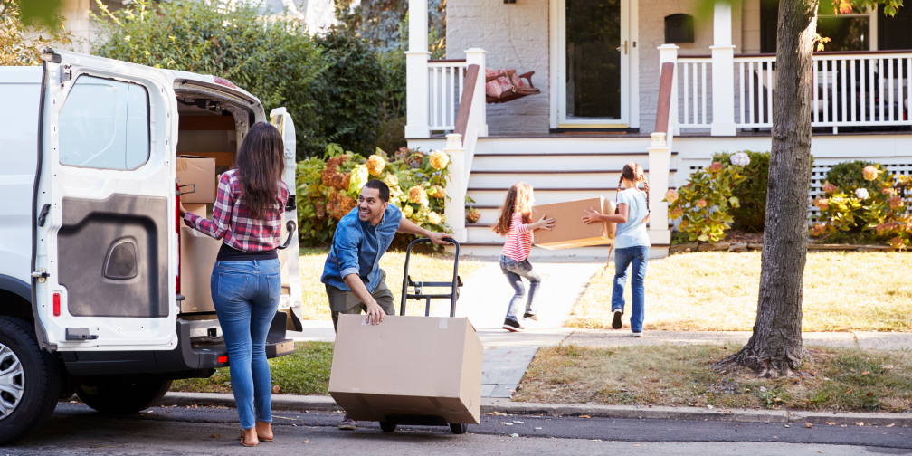 A family moving into a house. Two kids carry a box toward the house, a woman is about to remove a box from the back of a van filled with boxes and a man tilts a dolly with a box on it